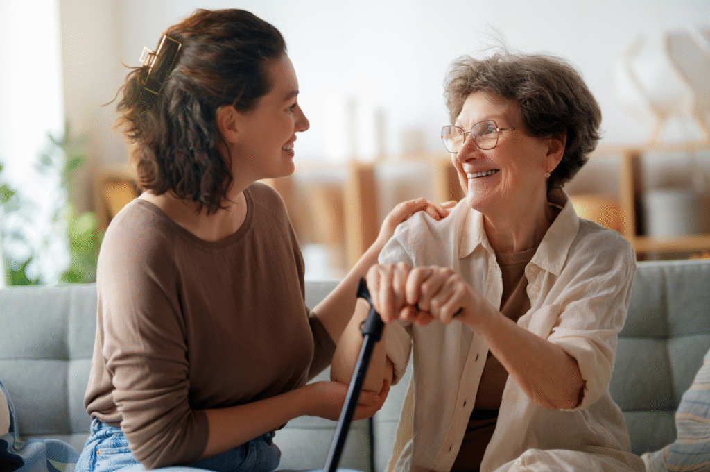 An elder person holsing a walking stick stitting next to a home care worker engaged in a pleasant conversation. Both are smiling.